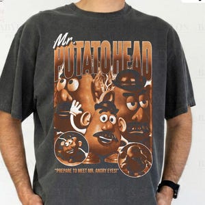 Può includere: T-shirt grigio scuro con una grafica di Mr. Potato Head in marrone e arancione, e la scritta "Mr. POTATOHEAD". La maglietta include anche il testo "PREPARE TO MEET MR. ANGRY EYES!" e "Comfort Colors Pepper".