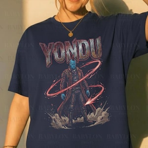 May include: Navy blue t-shirt featuring the word "YONDU" in a distressed font above a graphic of a blue-skinned character with a red arrow. The shirt has a crew neck and short sleeves. "Original Artist of This Design" is printed at the bottom.