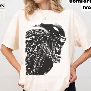 Puede incluir: Una camiseta de manga corta color marfil con un gran estampado gráfico negro de una criatura xenomorfa con la boca abierta y dientes visibles. La camiseta incluye el texto Babylon Prints y Comfort Colors Ivory en la esquina.