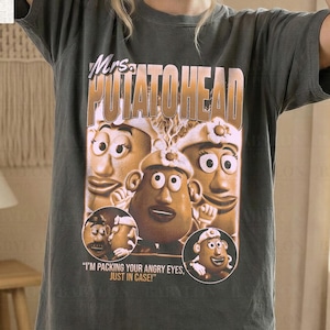 Può includere: T-shirt grigio scuro con una grafica dei personaggi di Mr. Potato Head e la scritta "Mrs. Potato Head". La maglietta ha anche la scritta "I'M PACKING YOUR ANGRY EYES, JUST IN CASE!". La maglietta è etichettata "Comfort Colors Pepper" e "Original Artist of This Design".