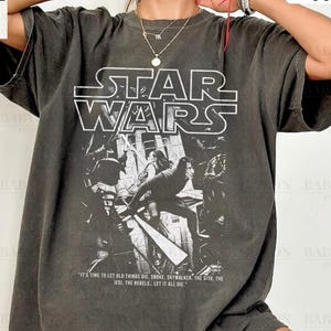 May include: A dark grey t-shirt with the Star Wars logo and a graphic of characters from the film. The shirt includes the text "It's time to let old things die..." and "Original Artist of This Design."