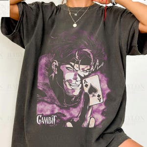 May include: A charcoal gray short-sleeve t-shirt featuring a graphic print of the character Gambit holding an ace of spades playing card. The design includes the text Gambit in a stylized font. The shirt is labeled as Comfort Colors Pepper.