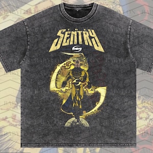 Puede incluir: Una camiseta gris oscuro de gran tamaño con un gráfico de superhéroe, la palabra "SENTRY" en letras doradas y el texto "Oversized Premium Tee!". La camiseta tiene un lavado de pimienta y es del artista original de este diseño.