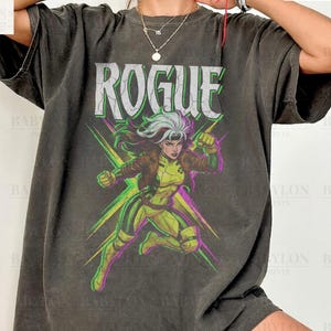 May include: A dark grey t-shirt with a graphic of the X-Men character Rogue. The word "ROGUE" is printed above the character in a bold, stylised font. The shirt also has the text "Comfort Colors Pepper" and "Original Artist of This Design".