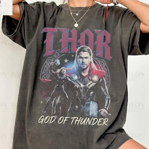 以下が含まれることがあります： 雷神ソーのグラフィックと赤い文字で「THOR」と書かれた濃い灰色のTシャツ。「GOD OF THUNDER」の文字と「Comfort Colors Pepper」の文字も含まれています。