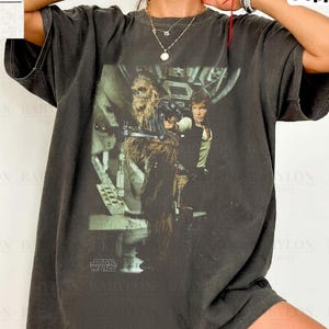 Könnte beinhalten: Ein anthrazitfarbenes Kurzarm-T-Shirt mit einem Grafikdruck von Chewbacca und Han Solo aus Star Wars. Das Shirt hat einen Rundhalsausschnitt und eine lockere Passform. Die Grafik zeigt die Figuren in einem Cockpit mit dem Star Wars-Logo.