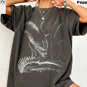 Puede incluir: Una camiseta de manga corta en color gris carbón con un estampado gráfico blanco de la criatura Xenomorfo de la franquicia Alien. La prenda tiene cuello redondo y un corte holgado, con el texto Comfort Colors Pepper impreso en la parte superior.