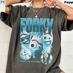 May include: Dark grey t-shirt featuring the word "FORKY" in large, teal letters above a graphic of three Forky characters. The shirt includes the text "I'M NOT A TOY. I'M A SPORK!" and is labeled "Comfort Colors Pepper."