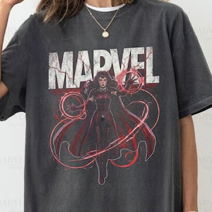 Puede incluir: Camiseta gris oscuro con el logo de Marvel y un gráfico rojo de un personaje con efectos de energía. La camiseta incluye el texto "Comfort Colors Pepper" y "Original Artist of This Design".