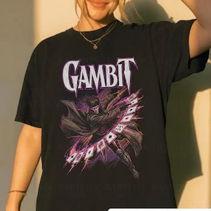 Pode incluir: Camiseta preta com a palavra "GAMBIT" em branco e um gráfico de um personagem jogando cartas de baralho. O design inclui detalhes em roxo e preto. A camisa tem o texto "Original Artist of This Design".