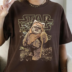 May include: Dark chocolate brown t-shirt featuring a Star Wars graphic. The design includes the Star Wars logo and an image of an Ewok character holding a staff. The shirt is from Babylon Prints.