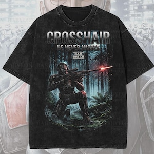 Könnte beinhalten: Schwarzes, übergroßes Premium-T-Shirt mit einer Grafik eines Charakters aus der Bad Batch-Serie, mit dem Text "CROSSHAIR HE NEVER MISSES". Das Design zeigt einen Soldaten, der in einer Waldumgebung mit einem Gewehr zielt. Das Shirt ist mit "Premium Wash: Black" gekennzeichnet.