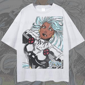 May include: An oversized white t-shirt featuring a graphic print of the comic book character Storm with flowing light blue hair, wearing a black and white costume with red circular accents. The word STORM is printed in blue at the bottom of the graphic.