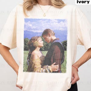 May include: Ivory-colored t-shirt featuring a romantic image of a couple in a field. The graphic is a vintage-style print. The shirt has short sleeves and the text "Comfort Colors Ivory" and "Original Artist of This Design".