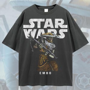 Puede incluir: Camiseta gris oscuro de gran tama&ntilde;o con el logotipo de Star Wars y una ilustraci&oacute;n del personaje Embo. El gr&aacute;fico es blanco, dorado y marr&oacute;n. El texto "EMBO" est&aacute; debajo del personaje. La camiseta tiene el texto "Oversized Tee!" y "Original Artist of This Design".