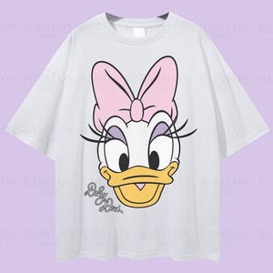 Puede incluir: Camiseta blanca de gran tama&ntilde;o con un dise&ntilde;o de Daisy Duck. El dise&ntilde;o incluye un lazo rosa, pesta&ntilde;as y el texto "Daisy Duck". La camiseta tiene mangas cortas y cuello redondo. El texto "Oversized White" est&aacute; en la parte superior.