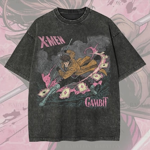 Pode incluir: Uma camiseta cinza escuro oversized com um gráfico do personagem Gambit dos X-Men. O design inclui o personagem em uma pose dinâmica com cartas de baralho e o texto "X-Men" e "Gambit". A camisa tem um acabamento "Premium Wash: Pepper".