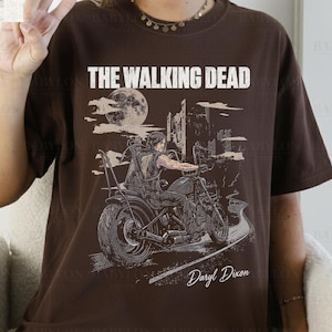 May include: Dark chocolate brown t-shirt featuring a graphic of a person riding a motorcycle with the text "THE WALKING DEAD" and "Daryl Dixon". The design includes a full moon and a cityscape. The shirt is from Babylon Prints.