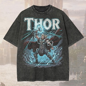 Könnte beinhalten: Ein dunkelgraues, übergroßes Premium-T-Shirt mit dem Wort "THOR" in Weiß über einer Grafik von Thor mit Hammer und Blitz. Das Shirt hat ein "Premium Wash: Pepper" Finish und den Text "Original Artist of This Design".
