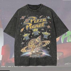 May include: A dark gray oversized t-shirt featuring a Pizza Planet graphic with aliens, a rocket, and planets. The shirt has the text "Pizza Planet" in yellow and blue. The shirt also has the text "Oversized Premium Tee!" and "Original Artist of This Design".