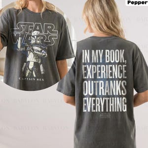 May include: A dark gray t-shirt featuring a Star Wars design. The front displays the Star Wars logo and a Captain Rex graphic. The back of the shirt has the text "IN MY BOOK, EXPERIENCE OUTRANKS EVERYTHING".