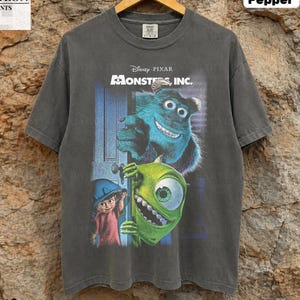 Puede incluir: Una camiseta de manga corta en color gris carbón con un estampado gráfico de los personajes Sulley, Mike Wazowski y Boo de Disney Pixar Monsters, Inc. La prenda tiene cuello redondo y se muestra en una percha de madera sobre un fondo de piedra.
