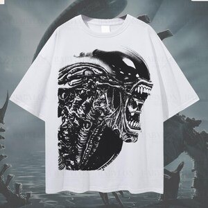 Puede incluir: Una camiseta blanca de corte oversize con un gran estampado gráfico en blanco y negro de una criatura biomecánica con cabeza alargada y dientes afilados. La prenda tiene mangas cortas y cuello redondo. Texto incluye Babylon Prints.
