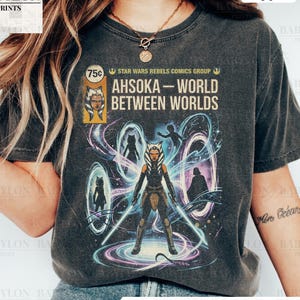 May include: Dark gray t-shirt featuring a graphic from Star Wars Rebels Comics Group. The design includes the text "AHSOKA - WORLD BETWEEN WORLDS" and an illustration of Ahsoka Tano with lightsabers. The shirt is labeled "Comfort Colors Pepper."