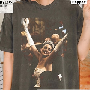 May include: A Comfort Colors pepper-coloured t-shirt with a vintage image of a woman in a gown, gloves, and tiara, arms raised. The shirt features the text "BABYLON PRINTS" and "Original Artist of This Design".