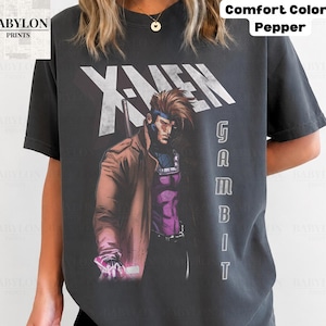 May include: A charcoal gray t-shirt featuring a graphic of the Marvel character Gambit from X-Men. The design includes the text X-MEN in large white letters above the character, who wears a brown trench coat and purple armor, with the word GAMBIT vertically.