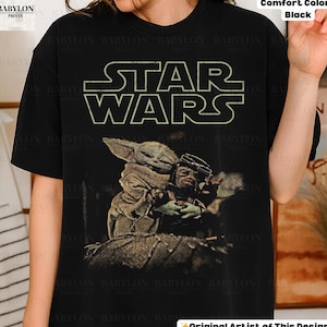 May include: Black t-shirt with the Star Wars logo in white, and a graphic of Grogu and a character. The shirt is labelled "Comfort Colors Black" and "Original Artist of This Design."