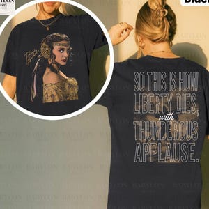 Puede incluir: Camiseta negra con un retrato de mujer con detalles dorados y el texto "Padmé Amidala" en dorado. La parte trasera de la camiseta muestra la cita "So this is how liberty dies, with thunderous applause."
