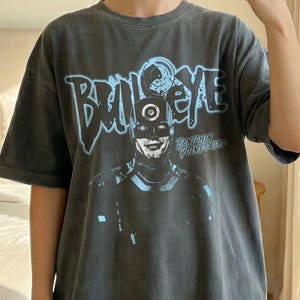 Könnte beinhalten: Dunkelgraues T-Shirt mit dem Wort "Bullseye" in Hellblau und einer Charakterillustration. Das Shirt trägt auch den Text "Comfort Colors Pepper" und "Original Artist of This Design". Model trägt Größe XL.