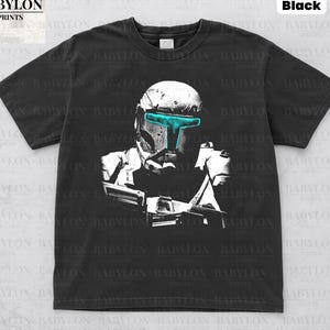 May include: Black t-shirt featuring a white and teal graphic of a helmeted character. The shirt has the text "Comfort Colors Black" at the top and "Original Artist of This Design" at the bottom. The Babylon Prints logo is in the upper left corner.