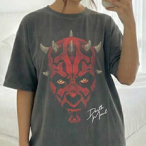 Może przedstawiać: Ciemnoszara koszulka z czerwono-czarną grafiką Darth Maula. Koszulka ma napis "Darth Maul" w białym skrypcie. Oznaczona jako "Comfort Colors Pepper" i "Original Artist of This Design". Modelka nosi rozmiar L.