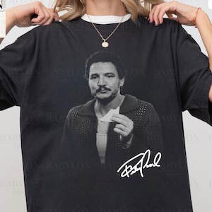 May include: Black Comfort Colors t-shirt featuring a black and white image of a person holding a card, with a signature below. The shirt has the text "Comfort Colors Black" and "Original Artist of This Design". The Babylon Prints logo is visible.