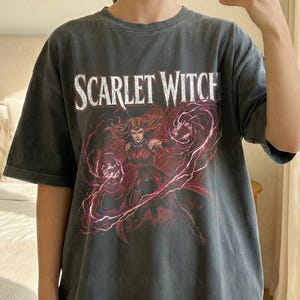 May include: A charcoal grey short-sleeve t-shirt featuring a graphic print of the Scarlet Witch with red energy swirls and the text "SCARLET WITCH" in white. The shirt is labelled as Comfort Colors Pepper and is shown in an XL size.