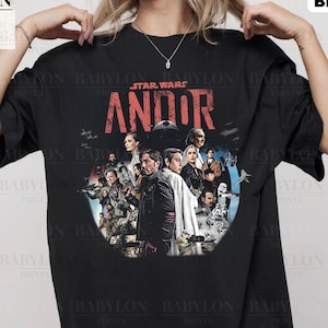 以下が含まれることがあります： 黒のTシャツに、スター・ウォーズ・アンドールのキャストのグラフィックと「スター・ウォーズ・アンドール」の赤い文字がプリントされています。モデルはXLサイズを着用しています。