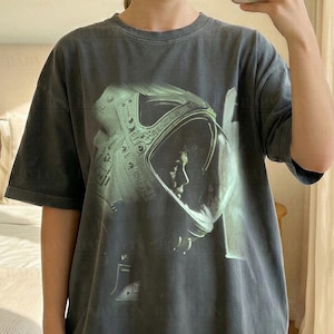 Può includere: Una t-shirt a maniche corte grigio antracite con una grande stampa grafica di un astronauta con casco. La maglietta è etichettata come Comfort Colors Pepper ed è di taglia XL. Il design include il testo Babylon Prints e Original Artist.