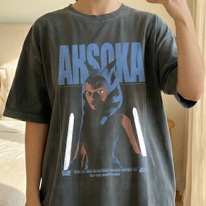 May include: Dark grey t-shirt featuring the word "AHSOKA" in blue block letters above a graphic of a character. The shirt also has the text "WHEN YOU FIND PEOPLE WHO NEED YOUR HELP, HELP THEM, NO MATTER WHAT". The shirt is a comfort color.