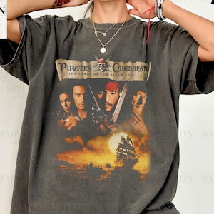 Op de afbeelding: Een donkergrijs T-shirt met een vintage-stijl afbeelding van de film Pirates of the Caribbean: The Curse of the Black Pearl. De afbeelding toont de hoofdpersonen uit de film, waaronder Captain Jack Sparrow, Will Turner en Elizabeth Swann.