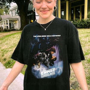 May include: A black t-shirt with a graphic print of a scene from the movie "Star Wars: The Empire Strikes Back". The text "The Star Wars Saga Continues" and "The Empire Strikes Back" are printed on the shirt.
