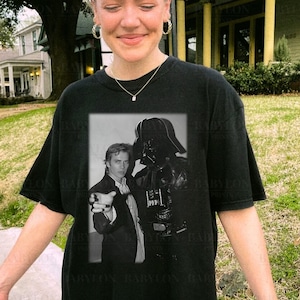 May include: A black t-shirt featuring a black and white image of Darth Vader and a man pointing at the viewer. The text "BABYLON PRINTS" is visible at the top of the image.