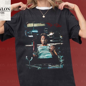 Könnte beinhalten: Ein schwarzes T-Shirt mit einem Grafikdruck von Robert Downey Jr. als Iron Man im Film Iron Man 3. Die Grafik zeigt Iron Man auf einem Stuhl sitzend, wobei ein rotes Licht von seinem Helm reflektiert wird. Der Text "Tony Stark" ist in Rot über der Grafik geschrieben.