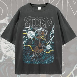 May include: Black oversized t-shirt featuring a graphic print of Storm, a superhero from Marvel Comics. The graphic depicts Storm in a superhero pose with lightning bolts surrounding her. The text "STORM" is printed above the graphic.