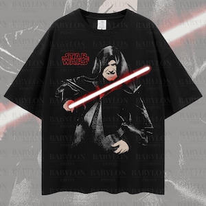 Puede incluir: Camiseta negra de gran tamaño con un estampado gráfico de un personaje de Star Wars sosteniendo un sable de luz. El texto "Star Wars" está impreso en la camiseta.
