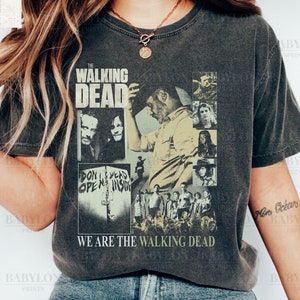 Puede incluir: Una camiseta gris oscuro con un estampado gráfico desgastado de personajes de la serie de televisión "The Walking Dead". El gráfico incluye el título de la serie, un grupo de personajes y el texto "We are the Walking Dead".