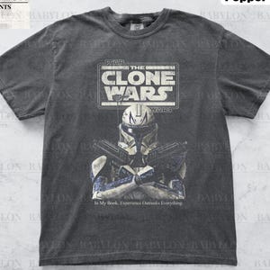 Pode incluir: Uma camiseta cinza escura com um visual desbotado apresentando um gráfico de um personagem de Star Wars Clone Wars, um capacete branco com marcas azuis, segurando dois blasters. O texto "STAR THE CLONE WARS" está acima do personagem e "WARS" está abaixo do personagem. O texto "In My Book, Experience Outranks Everything." está abaixo do personagem.