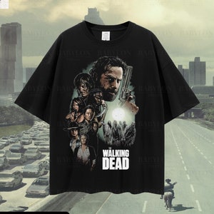 Puede incluir: Camiseta negra con un estampado gráfico de personajes de la serie de televisión "The Walking Dead". El gráfico presenta a Rick Grimes, Daryl Dixon, Michonne y otros personajes de la serie. El texto "The Walking Dead" está impreso debajo del gráfico.