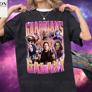 Könnte beinhalten: Ein schwarzes T-Shirt mit einem Grafikdruck der Guardians of the Galaxy-Charaktere. Die Charaktere sind im Retro-Stil mit einem rosa und violetten Farbschema gehalten. Der Text "Guardians of the Galaxy" ist in rosa und violetten Buchstaben gedruckt.